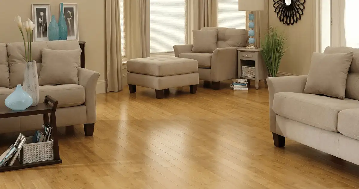 A living room with professionally installed bamboo flooring, beige sofas, a matching ottoman, a side table with a blue lamp, vases, framed pictures, and large windows with blinds and curtains in San Jose, CA
